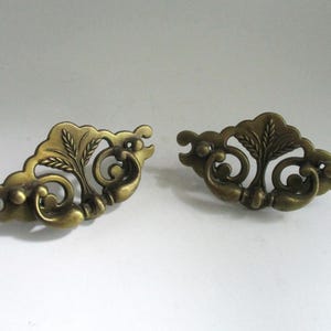 May include: Two antique brass drawer pulls with an ornate, floral design. Each pull features a stylized leaf and scroll pattern, with a warm, aged finish. These decorative hardware pieces are ideal for vintage furniture restoration.