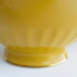 May include: A close-up of a yellow, vintage-style bowl. The bowl has a rounded shape with vertical ridges on the sides and a small, circular base. The base is trimmed with a gold-coloured band.
