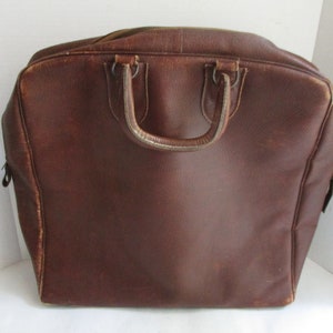 May include: A brown leather travel bag with a top handle and a zipper closure. The bag has a rectangular shape with rounded corners and a worn appearance. The handle is attached with metal rings.