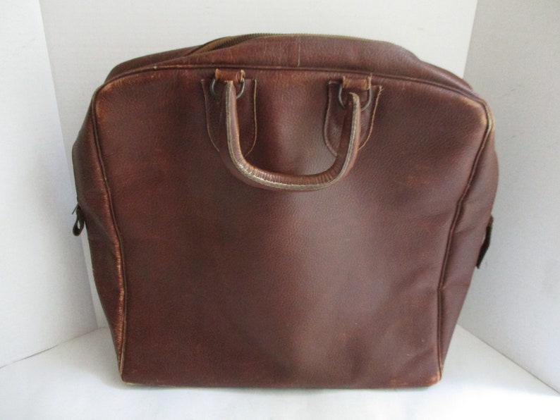 May include: A brown leather travel bag with a top handle and a zipper closure. The bag has a rectangular shape with rounded corners and a worn appearance. The handle is attached with metal rings.