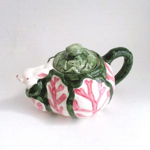 May include: A small, ceramic teapot with a white base, adorned with pink coral-like patterns and green accents. The lid and handle are also green. The teapot has a small spout and a rounded shape, perfect for serving tea.
