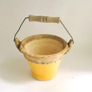 May include: A small, yellow ceramic bucket with a light brown rim and a wooden handle. The handle is attached to the bucket with a wire. The bucket is empty and sits on a white surface.