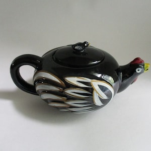 May include: Black ceramic teapot with a chicken design. The teapot features a black handle and spout, with white, gold, and silver feather details. The lid has a small bird figurine. The chicken's head is red, yellow, and white.