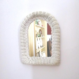 May include: White wicker arched mirror. The mirror is mounted on a white wall. The reflection shows a glimpse of a room with shelves and various decorative items.