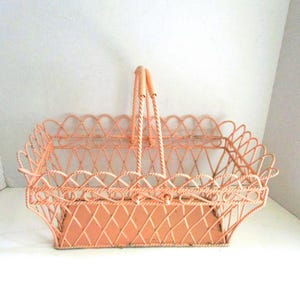 May include: A peach-colored metal basket with a handle. The basket has a rectangular shape with a scalloped edge and a woven design. The handle is made of twisted rope and is attached to the basket with metal loops.
