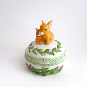 May include: A decorative ceramic trinket box, egg-shaped, with a lid. The box is white with green and pink floral accents. Two orange rabbits sit atop the lid. The box is likely used for storing small jewelry items.