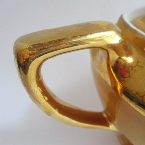 May include: Close-up of a gold-coloured handle of a small ceramic container. The handle is curved and has a glossy finish with some wear. The container appears to be part of a tea set or similar serving item.