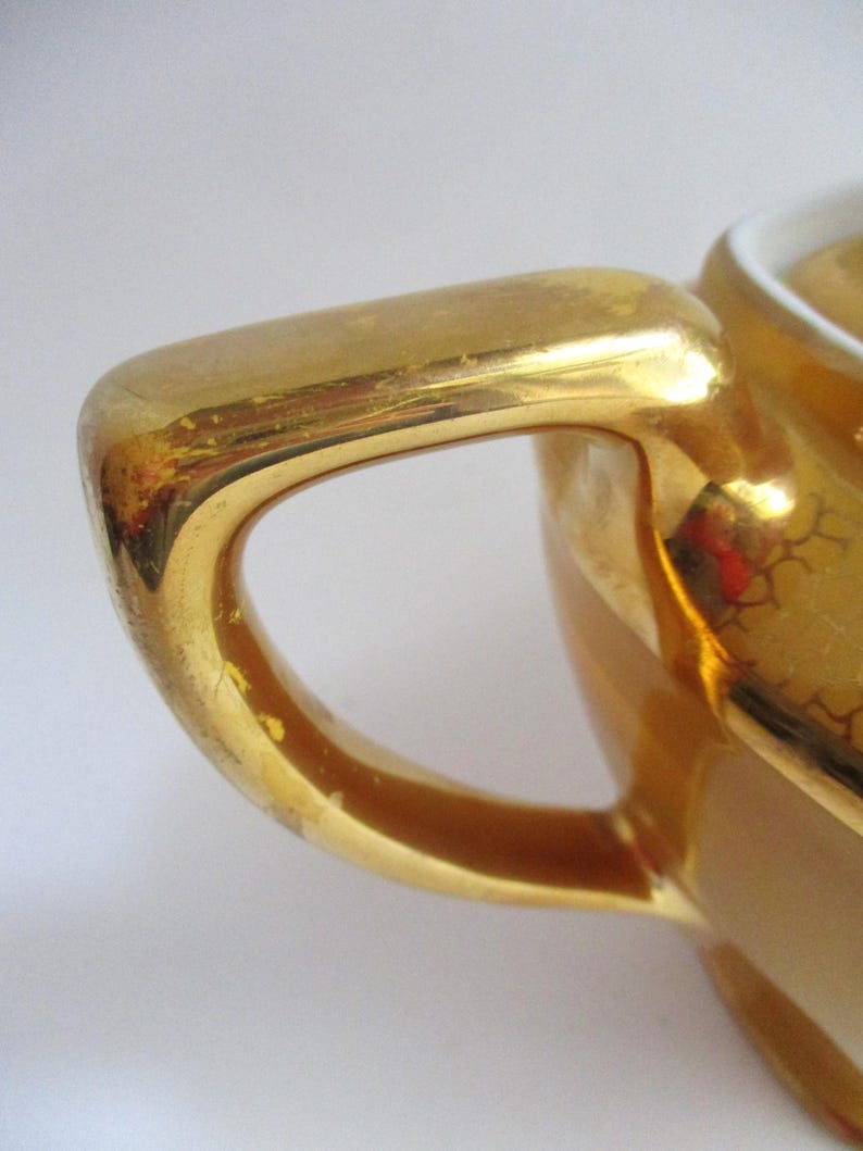 May include: Close-up of a gold-coloured handle of a small ceramic container. The handle is curved and has a glossy finish with some wear. The container appears to be part of a tea set or similar serving item.