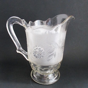 May include: Clear glass pitcher with a decorative handle and a scalloped rim. The pitcher features a textured band with floral and geometric patterns. The base is a pedestal design.