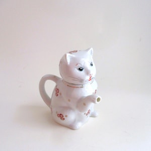 May include: A white ceramic teapot in the shape of a cat. The teapot has a handle, a spout, and is decorated with small red floral patterns. The cat's face has blue eyes and a red nose. The teapot is a decorative item.