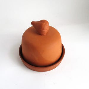 May include: A terracotta-colored butter dish with a rounded lid and a small, decorative handle. The dish sits on a matching round base. The design is simple and functional, suitable for kitchen use.