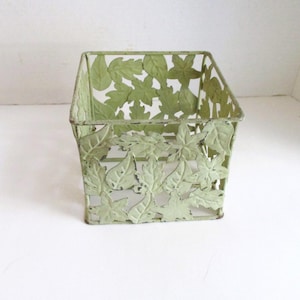 May include: A square, light green metal basket with a leaf design. The basket has cut-out leaves on the sides and is slightly distressed. The basket is empty and sits on a white surface. Ideal for home decor.