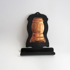 May include: Black metal wall mounted holder with a copper-colored stove design. The holder has a small shelf below the stove design.