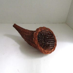 May include: A brown woven wicker cornucopia basket featuring a tapered, curved horn shape and a braided rim. The basket is constructed from thin, flexible reeds woven into a tight, sturdy pattern that narrows to a point at the closed end.
