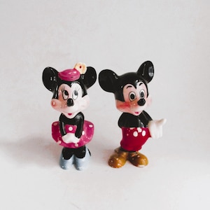 May include: A pair of ceramic figurines of Mickey Mouse and Minnie Mouse. Mickey is wearing red shorts with white polka dots and white gloves. Minnie is wearing a pink dress with white polka dots and a pink bow. Both figurines have black ears and black shoes.
