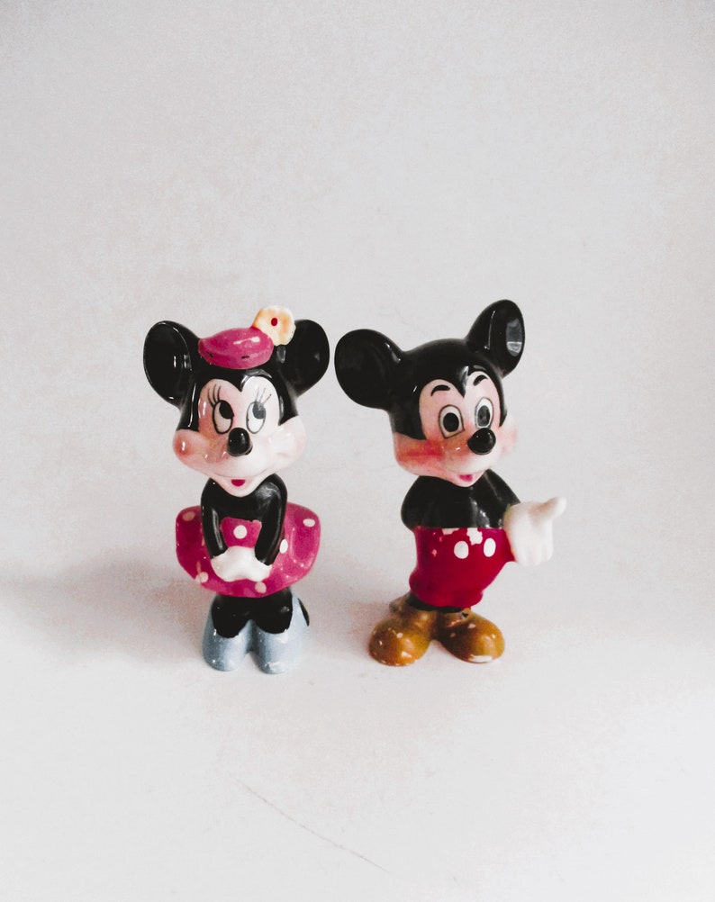 May include: A pair of ceramic figurines of Mickey Mouse and Minnie Mouse. Mickey is wearing red shorts with white polka dots and white gloves. Minnie is wearing a pink dress with white polka dots and a pink bow. Both figurines have black ears and black shoes.