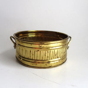 May include: A gold-toned metal planter with two handles. The planter has a ribbed design and is oval shaped.