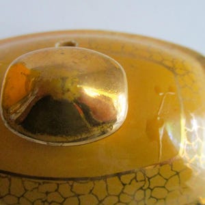 May include: Close-up of a gold-coloured lid with a rounded knob. The lid is a warm yellow with a decorative pattern in a darker gold colour. The knob reflects the surrounding environment.