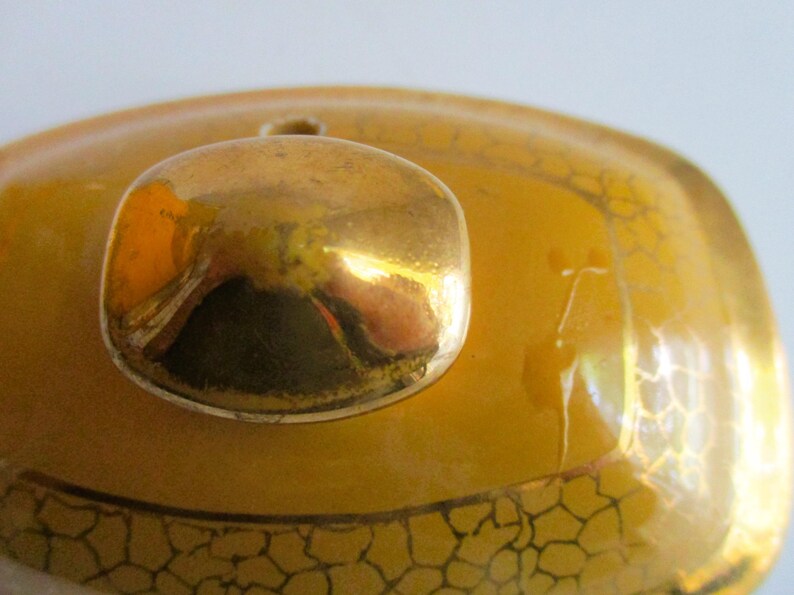 May include: Close-up of a gold-coloured lid with a rounded knob. The lid is a warm yellow with a decorative pattern in a darker gold colour. The knob reflects the surrounding environment.