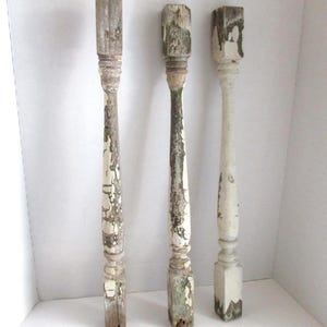 May include: Three weathered wooden balusters with peeling white paint and distressed details. The balusters have a rectangular top and bottom with a turned, decorative center section. The wood shows signs of age and exposure, adding to their rustic charm.