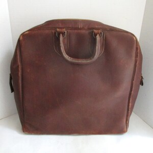 May include: A brown leather briefcase with a top handle and a rectangular shape. The bag has a zipper closure and appears to be vintage. The leather has a worn appearance, suggesting age and use. The briefcase is designed for carrying documents and other essentials.