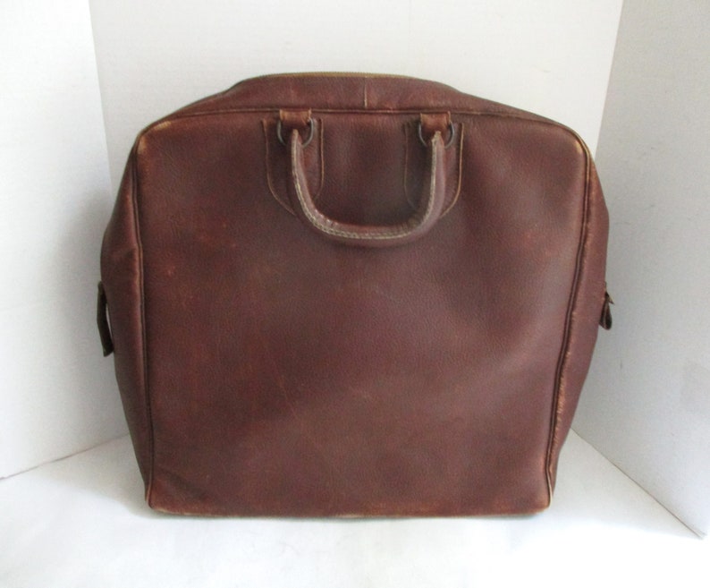 May include: A brown leather briefcase with a top handle and a rectangular shape. The bag has a zipper closure and appears to be vintage. The leather has a worn appearance, suggesting age and use. The briefcase is designed for carrying documents and other essentials.