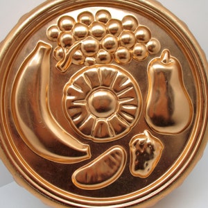 Puede incluir: Un molde para pastel de color cobre con un dise&ntilde;o en relieve de un pl&aacute;tano, pi&ntilde;a, pera y uvas. El molde es redondo y tiene un borde acanalado.