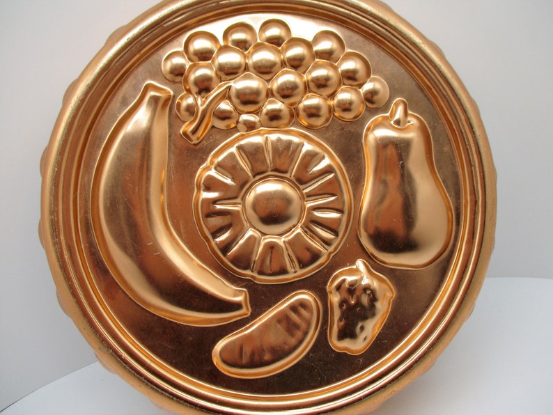 Puede incluir: Un molde para pastel de color cobre con un dise&ntilde;o en relieve de un pl&aacute;tano, pi&ntilde;a, pera y uvas. El molde es redondo y tiene un borde acanalado.