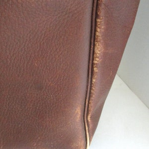 May include: Close-up of a brown leather bag. The leather has a textured surface with visible grain and slight imperfections. The edges of the bag show wear and a lighter color, indicating age and use. The stitching is visible along the seams.