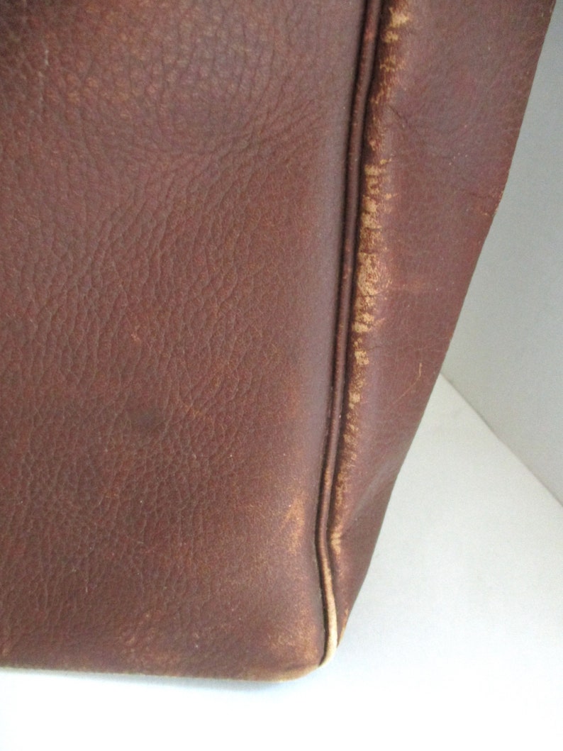 May include: Close-up of a brown leather bag. The leather has a textured surface with visible grain and slight imperfections. The edges of the bag show wear and a lighter color, indicating age and use. The stitching is visible along the seams.