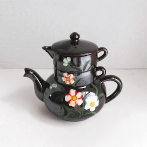 May include: A black ceramic teapot with a floral design. The teapot has a lid and a spout. The flowers are painted in shades of pink, white, and green.