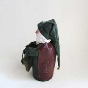 May include: A green and red Santa Claus figurine with a white beard and a green hat with a gold bell. He is holding a green sack in his left hand.