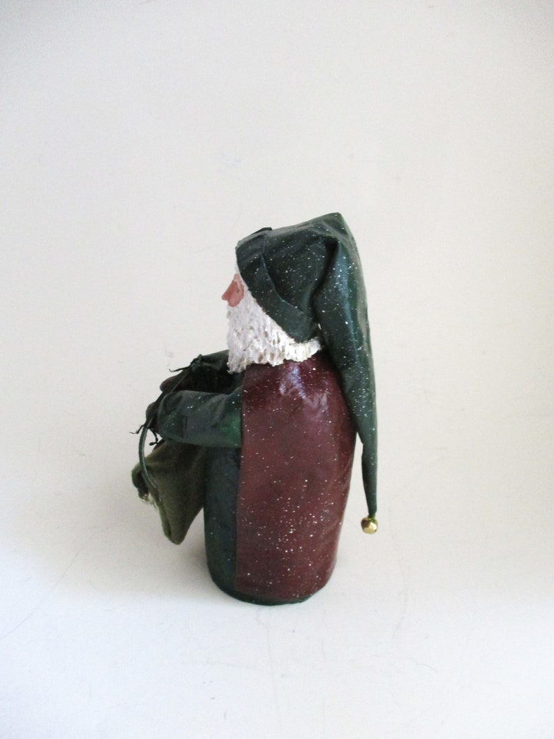 May include: A green and red Santa Claus figurine with a white beard and a green hat with a gold bell. He is holding a green sack in his left hand.