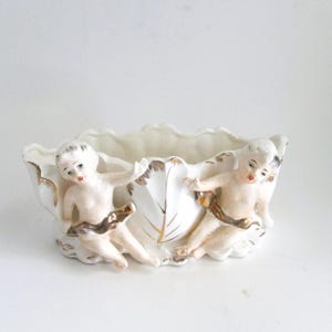 May include: A white ceramic bowl with a scalloped rim, featuring two cherubic figures with gold accents. The bowl is shaped like a leaf, with gold detailing on the edges and the central vein.
