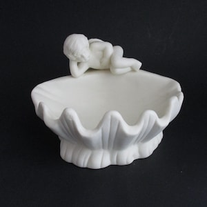 May include: A white ceramic seashell-shaped dish with a reclining cherub figurine. The dish has a scalloped edge and a textured base. The cherub is lying on its side, resting its head on its hand.