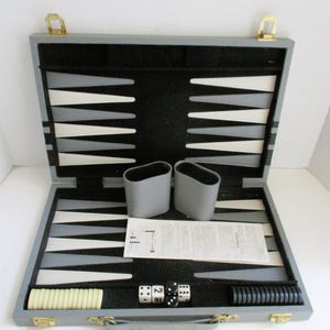 May include: Open backgammon set with gray case, black and white playing surface, and accessories. Includes dice, dice cups, and playing pieces. The case has gold-colored clasps and hinges.