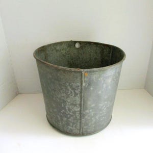 May include: A galvanized metal bucket with a slightly tapered cylindrical shape. The bucket has a weathered, mottled gray appearance with a rolled rim and a small hole near the top. It is likely used for storage or decorative purposes.