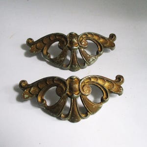 May include: Two matching metal drawer pulls featuring an ornate, symmetrical scrollwork design with a weathered gold-tone finish. Each piece has a central raised element and two open, curved cutouts, showing signs of patina and surface wear.