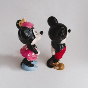 May include: A pair of ceramic figurines of Mickey Mouse and Minnie Mouse. Mickey is wearing red shorts and yellow shoes. Minnie is wearing a pink and white polka dot dress and blue shoes.