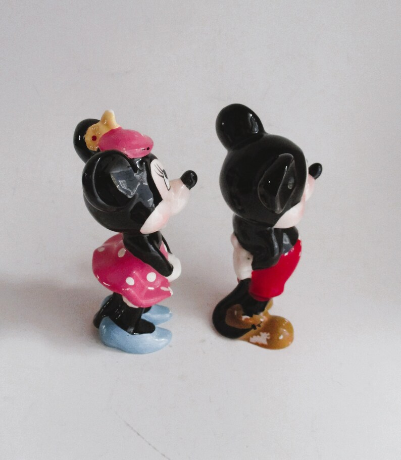 May include: A pair of ceramic figurines of Mickey Mouse and Minnie Mouse. Mickey is wearing red shorts and yellow shoes. Minnie is wearing a pink and white polka dot dress and blue shoes.