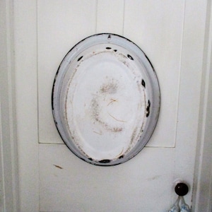May include: A white oval-shaped metal tray with a black rim hangs on a white wooden door. The tray has a worn, vintage look.