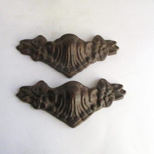 May include: Two dark brown metal furniture corner accents featuring a raised, symmetrical relief pattern. Each piece has a central pointed arch design flanked by floral motifs on both sides, with a textured, weathered finish throughout the surface.