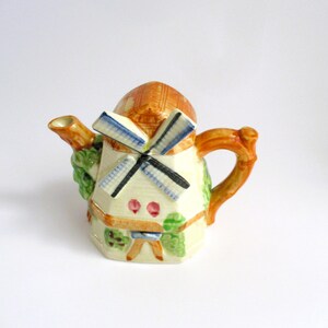 May include: A small ceramic teapot shaped like a windmill with a brown roof, white walls, blue and black windmill blades, and green and brown painted details.
