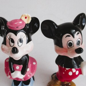 May include: A pair of ceramic figurines of Mickey Mouse and Minnie Mouse. Mickey is wearing red shorts with white polka dots and a white glove. Minnie is wearing a pink dress with white polka dots and a pink hat with a yellow flower.