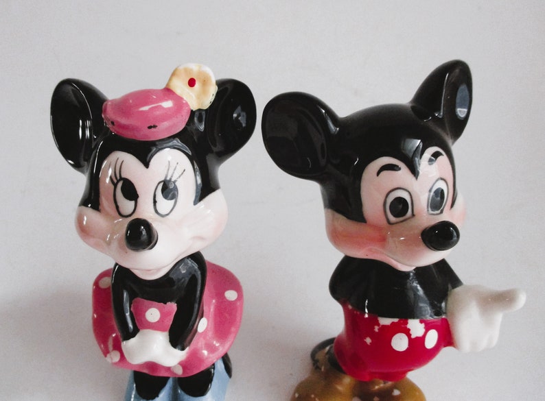 May include: A pair of ceramic figurines of Mickey Mouse and Minnie Mouse. Mickey is wearing red shorts with white polka dots and a white glove. Minnie is wearing a pink dress with white polka dots and a pink hat with a yellow flower.