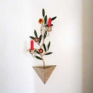 May include: A decorative wall sconce with a floral and leaf design. The sconce features two red candles and is made of metal. The sconce has a triangular base and is mounted on a white wall. The flowers are orange and pink.