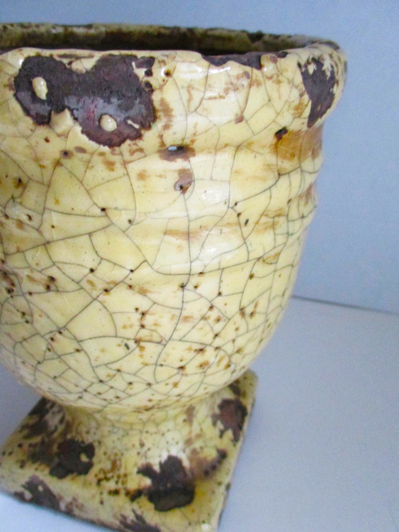 May include: A yellow ceramic planter with a square base and a cracked glaze. The planter has a distressed look with brown and white markings.