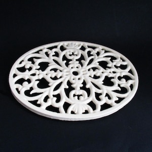 May include: A white, oval-shaped trivet with an intricate, openwork design. The trivet features a symmetrical pattern of swirling lines and floral motifs, creating a delicate and ornate appearance. The trivet is made of a solid material and is set against a dark background.