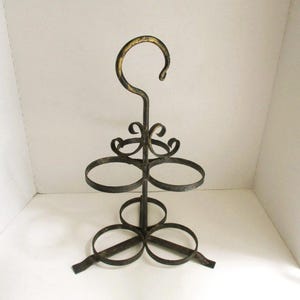 May include: A wrought iron wine bottle holder with a hook at the top and three circular holders. The metal is dark with gold accents, and the design includes decorative swirls. The stand is designed to hold multiple bottles.