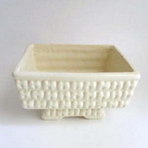 May include: Off-white ceramic planter with a rectangular shape and a textured basket weave design. The planter sits on small, rounded feet. The interior is a lighter shade of off-white.